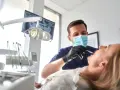 Dentysta w masce wykonuje szynowanie zębów pacjentce. W tle widać aparat RTG i lampę zabiegową.