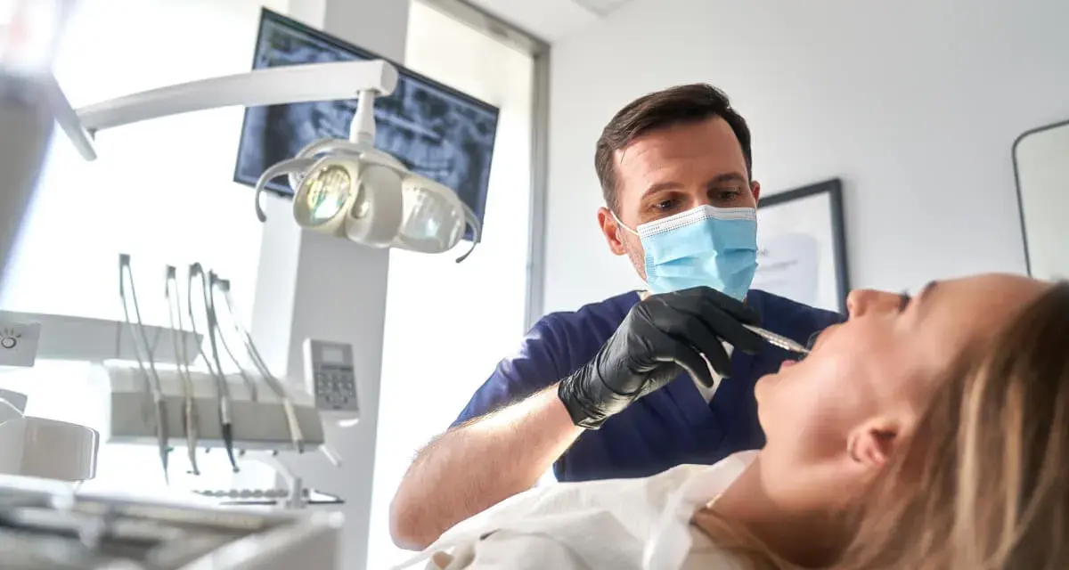 Dentysta w masce wykonuje szynowanie zębów pacjentce. W tle widać aparat RTG i lampę zabiegową.