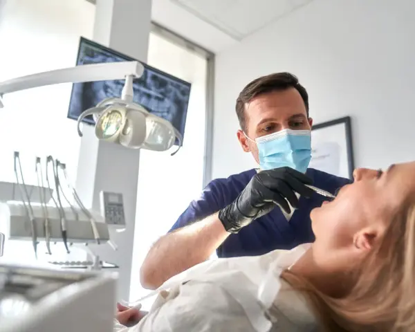 Dentysta w masce wykonuje szynowanie zębów pacjentce. W tle widać aparat RTG i lampę zabiegową.