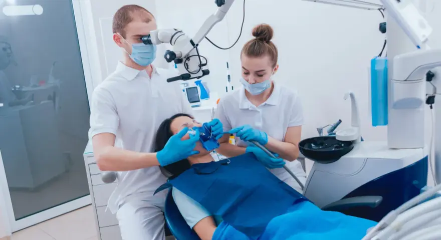 Dentysta pod mikroskopem bada ząb pacjentki, by wykluczyć zatrucie zęba. Asystentka przygotowuje narzędzia.