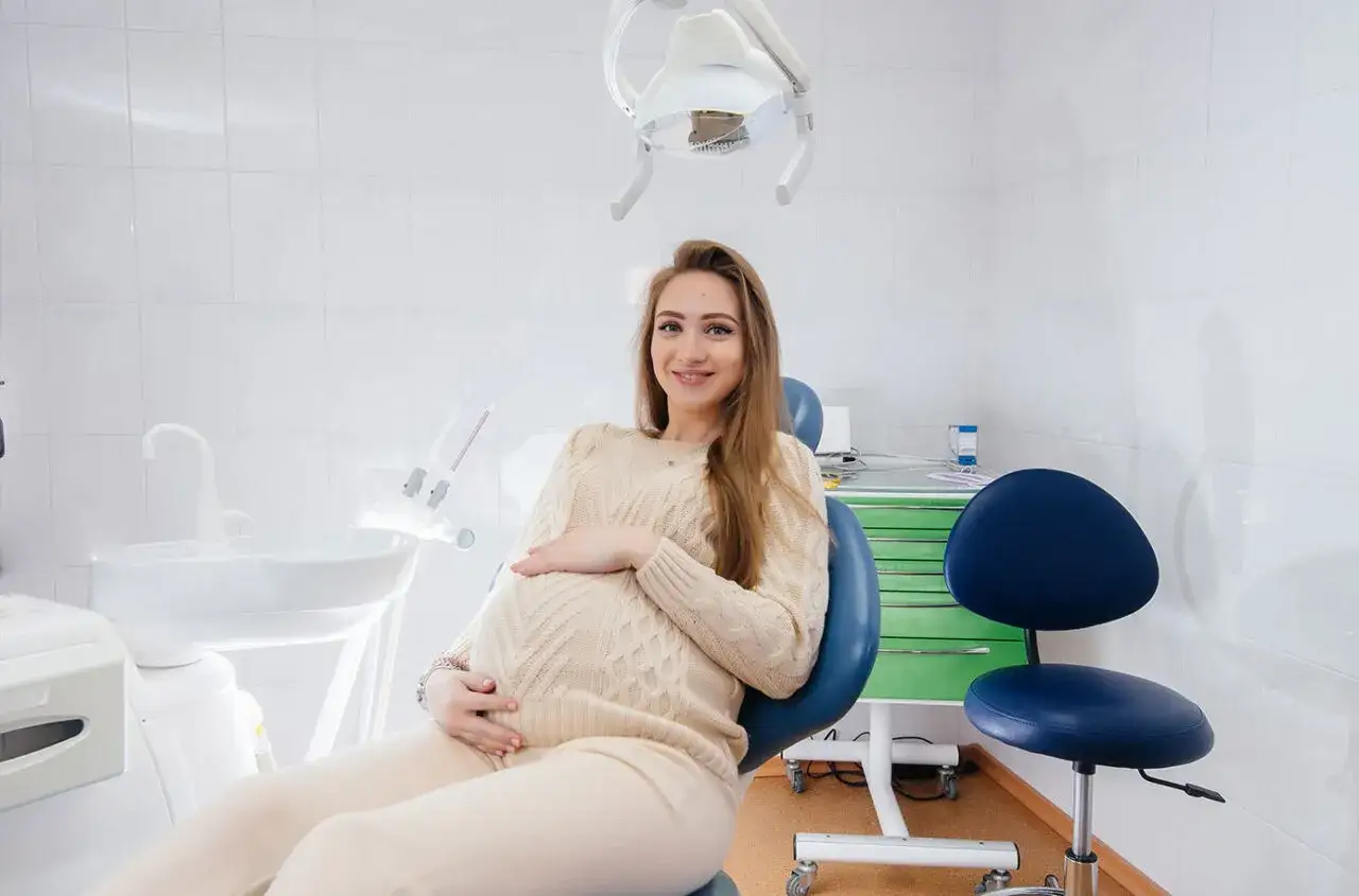 Kobieta w 3 trymestrze ciąży, z dłonią na brzuchu, siedzi na fotelu dentystycznym. Może to być wizyta związana z bólem zęba w ciąży.