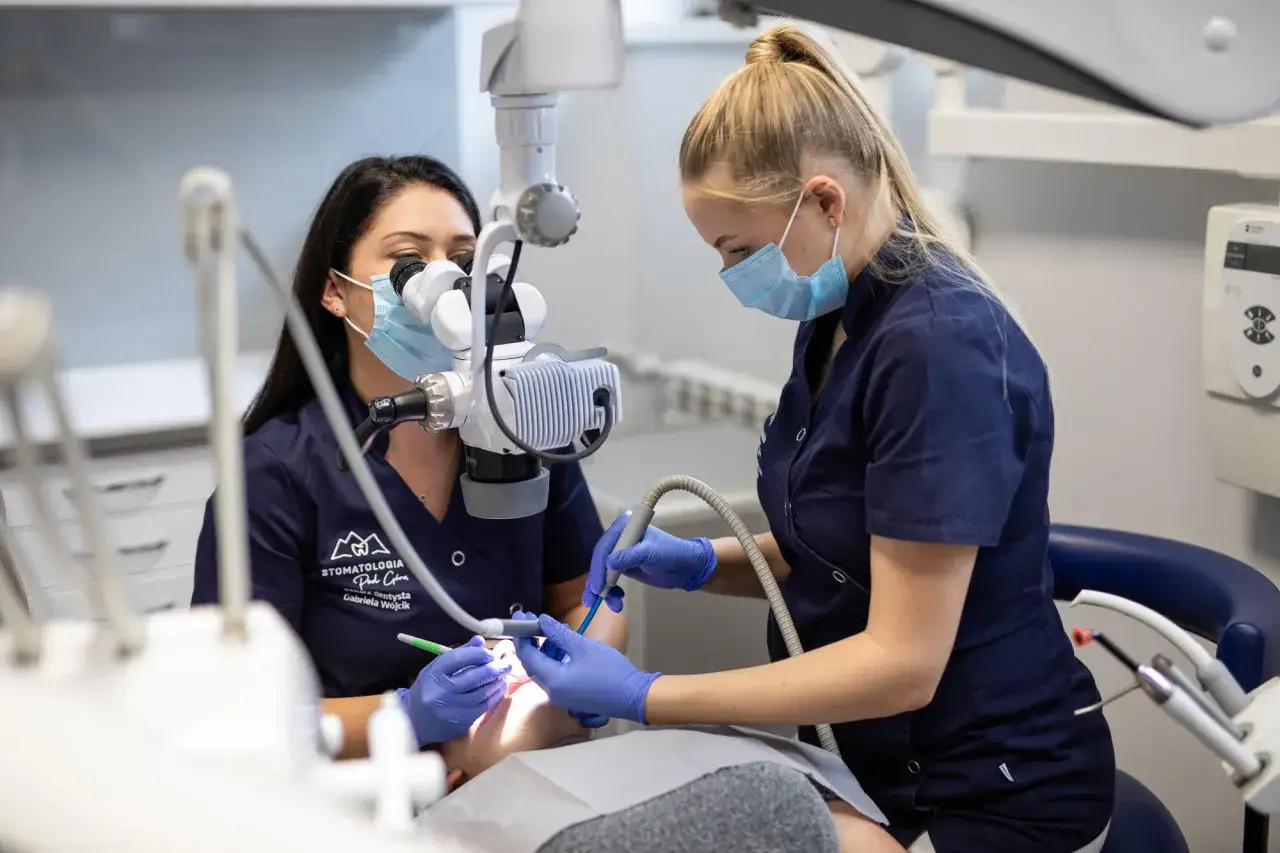 Dwie dentystki w maseczkach pracują przy pacjencie. Jedna używa mikroskopu, druga wierci. Nawet w środku nocy, gdy ząb boli, można liczyć na pomoc.